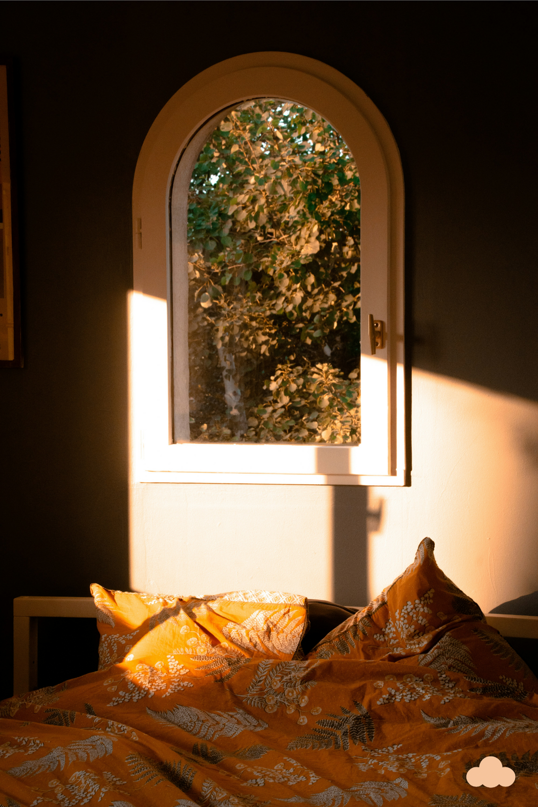 Sunlight pours through an arched window, casting a warm glow over a cozy bed with orange botanical-patterned bedding, creating a serene and inviting atmosphere.